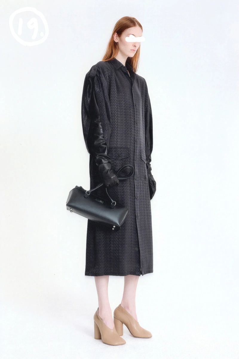 A slender fashion model stands in a studio, wearing a long patterned coat and black leather gloves, holding a structured black leather handbag.