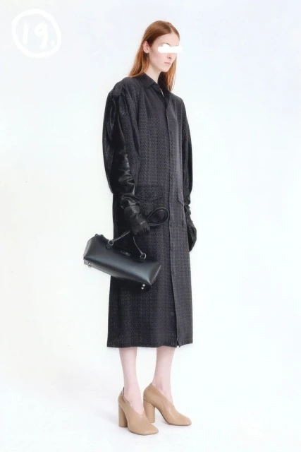 A slender fashion model stands in a studio, wearing a long patterned coat and black leather gloves, holding a structured black leather handbag.