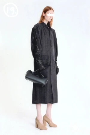 Model in dark patterned coat holding a black handbag against a white background