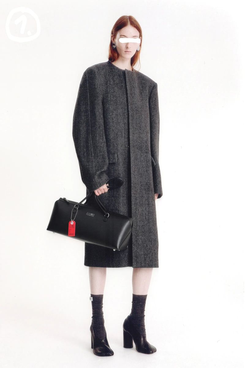 Fashion model poses in a long gray coat, holding a black duffel bag, with a white studio backdrop. High-fashion, minimalist aesthetic.