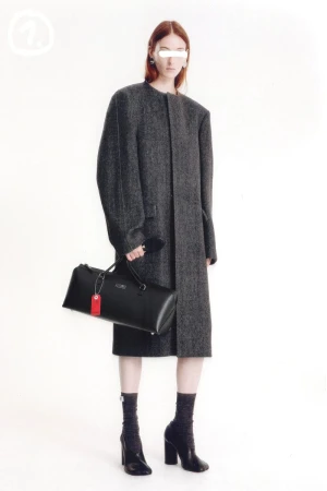 Fashion model poses in a long gray coat, holding a black duffel bag, with a white studio backdrop. High-fashion, minimalist aesthetic.