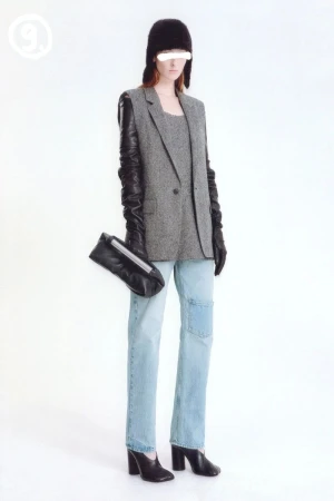 Fashionable woman in light-wash jeans, grey blazer, and black leather accessories poses against a bright white backdrop in a studio shot.