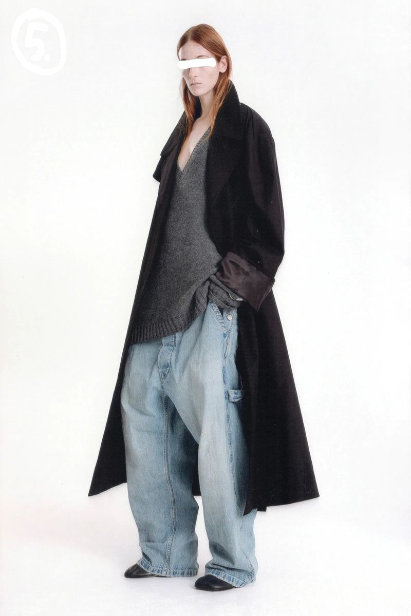 Adult female fashion model in a black coat and oversized light-wash denim jeans, with a grey v-neck sweater, standing in a studio.