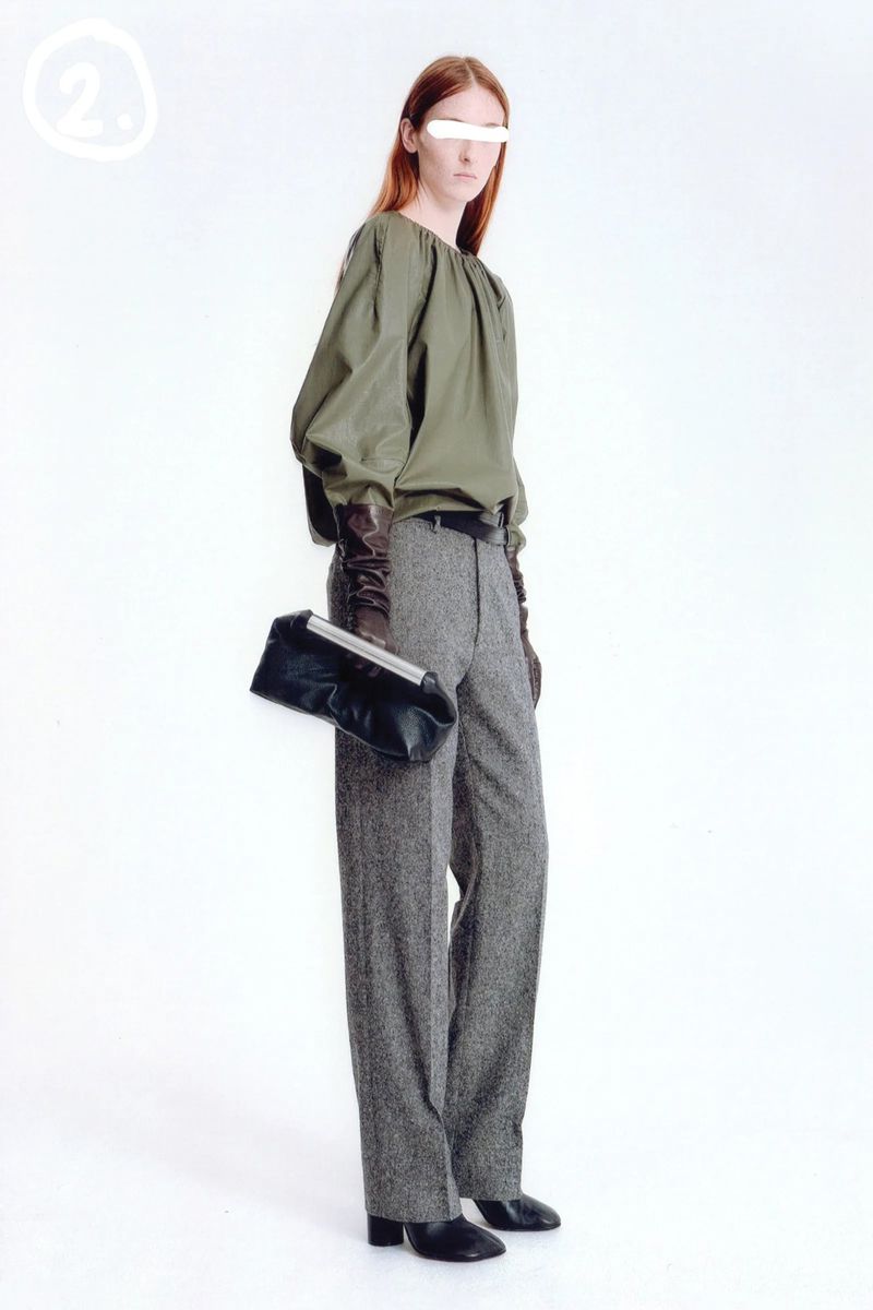 Fashion model poses in an olive blouse and tweed pants, holding a clutch purse in a bright studio setting with a modern aesthetic.