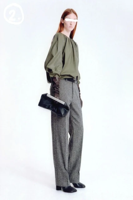 Fashion model poses in an olive blouse and tweed pants, holding a clutch purse in a bright studio setting with a modern aesthetic.