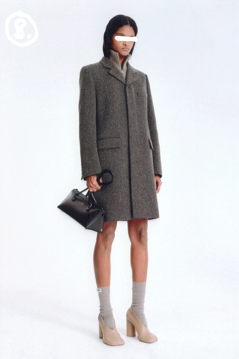 Woman in a gray coat holding a black purse stands in a white studio wearing beige socks and heels for a fashion editorial style shoot.