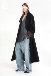 Model wearing a long black coat, grey V-neck sweater, and baggy light blue jeans against a white background