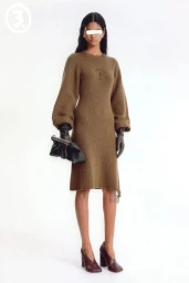 Model wearing a brown knit dress, dark gloves, and high heels, holding a black clutch against a white seamless backdrop.