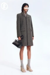 Model standing against a plain white background wearing a tweed coat, grey socks, and heels, holding a black handbag.