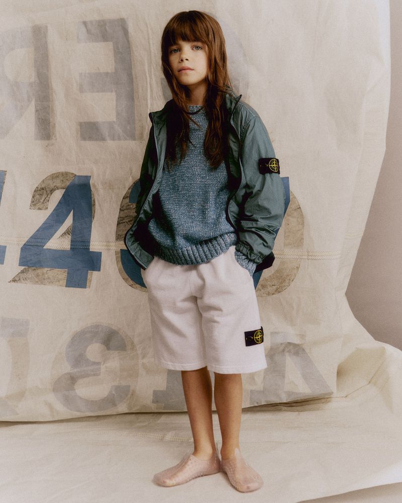 Young child poses in an outdoor setting, wearing a blue sweater, windbreaker jacket, white shorts, and ballet flats with a light background.