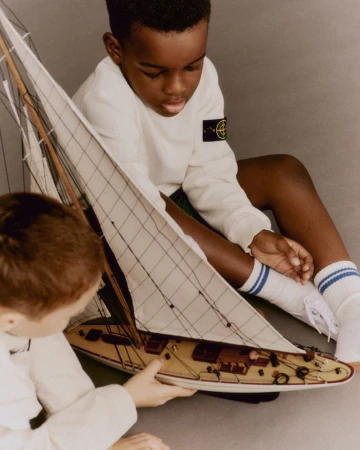 Two children playing with a model sailboat indoors in a close-up shot, showcasing a fashion advertisement with a neutral backdrop.