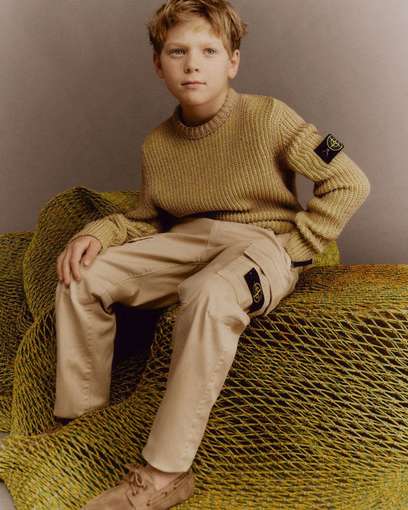 A young boy sits with a neutral expression wearing a knit sweater, cargo pants, and suede shoes in a studio setting.