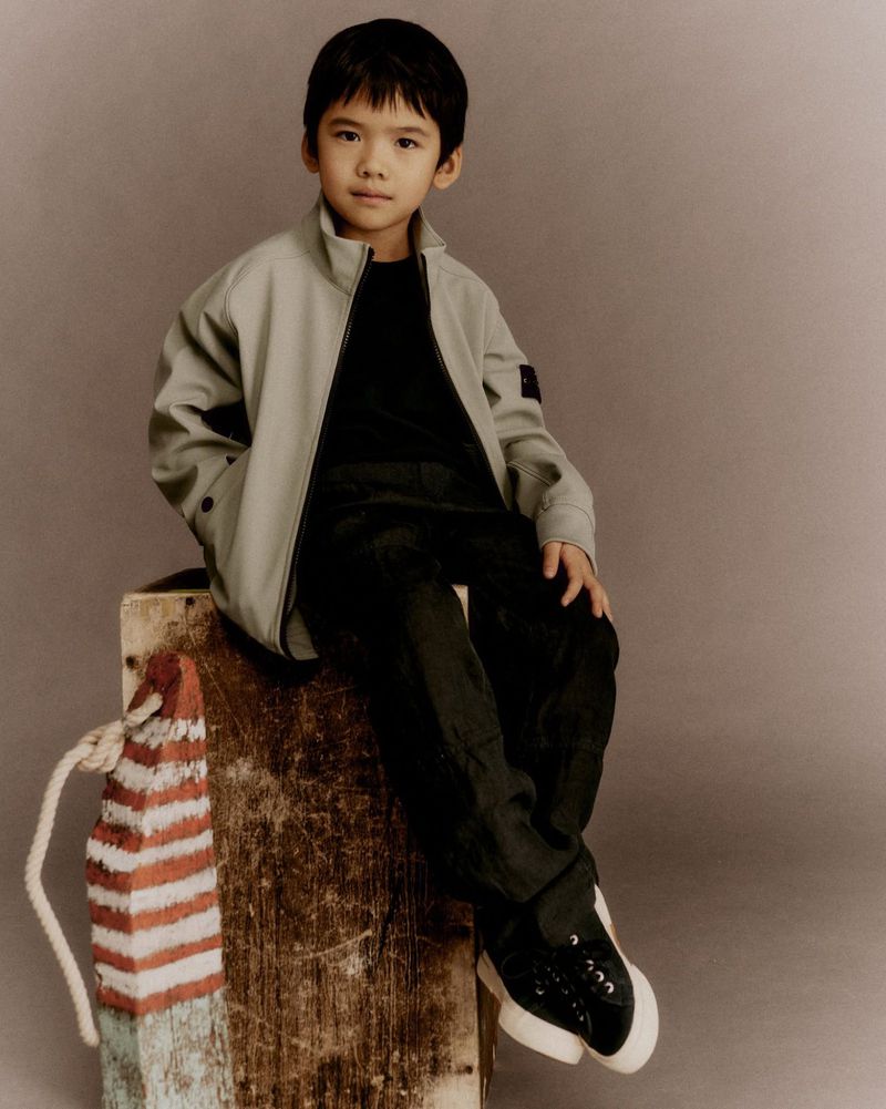 Young boy in a gray jacket and black pants sits on a wooden crate with a painted bollard prop; studio shot with a neutral background.