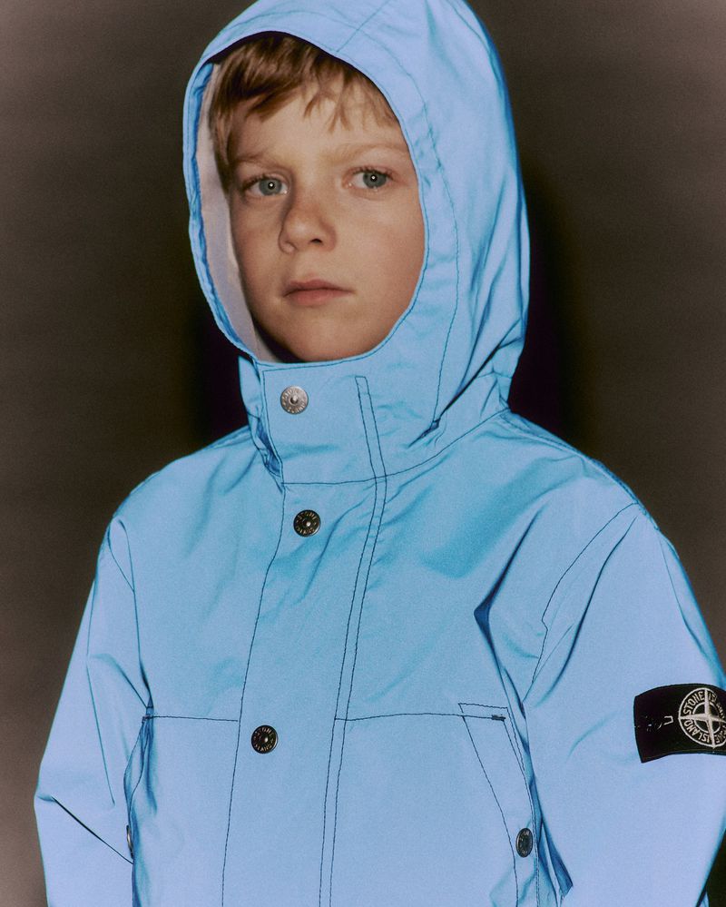 Boy wearing a light blue hooded jacket with buttons and a black Stone Island logo patch on his left arm in a studio setting.