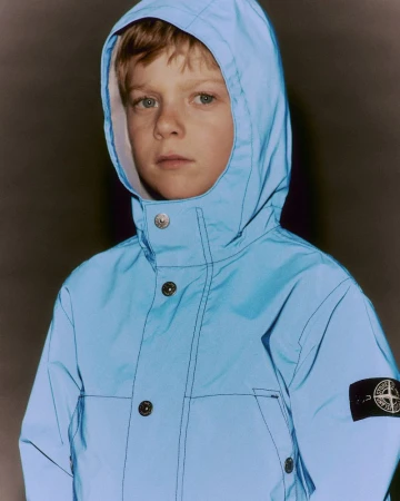Boy wearing a light blue hooded jacket with buttons and a black Stone Island logo patch on his left arm in a studio setting.