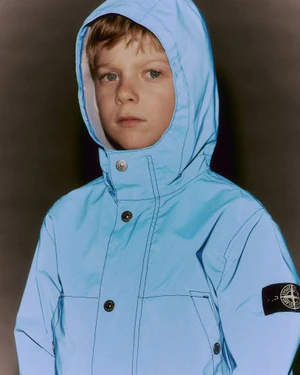 Child wearing a light blue hooded jacket with a compass logo patch on the sleeve against a dark neutral studio background.