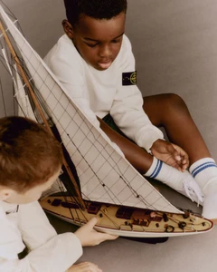 Two young boys interacting with a detailed model sailboat in a minimalist studio setting for Stone Island.