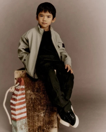 Young child model wearing a light grey jacket and black pants sitting on a wooden crate for Stone Island Junior SS26.