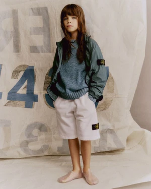 Young model wearing a green windbreaker, blue sweater, and white shorts standing against a sail backdrop