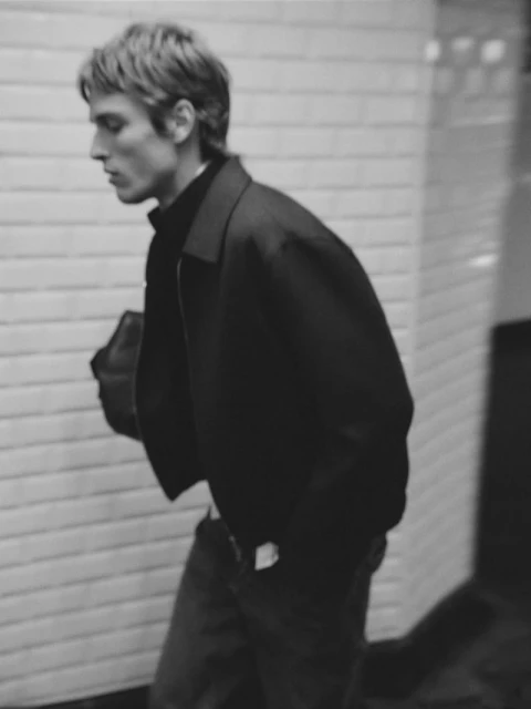 Monochrome shot of a blonde adult male in a black jacket and grey pants walking in an indoor, tiled environment, side profile.