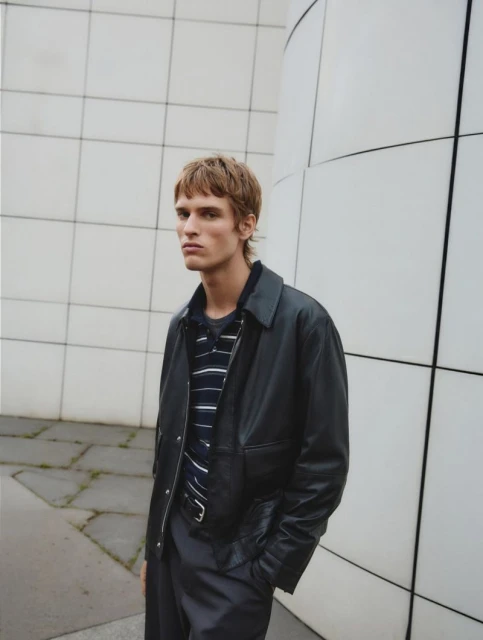 Male model poses in a black leather jacket and striped shirt in front of a grey tiled wall, creating a fashion editorial image.