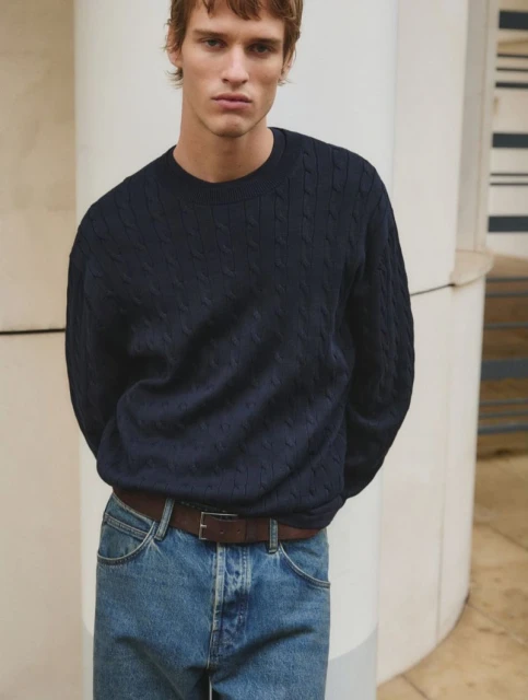 A fair-skinned adult male poses in a navy cable-knit sweater, light blue jeans, and a brown belt against a neutral, architectural backdrop.