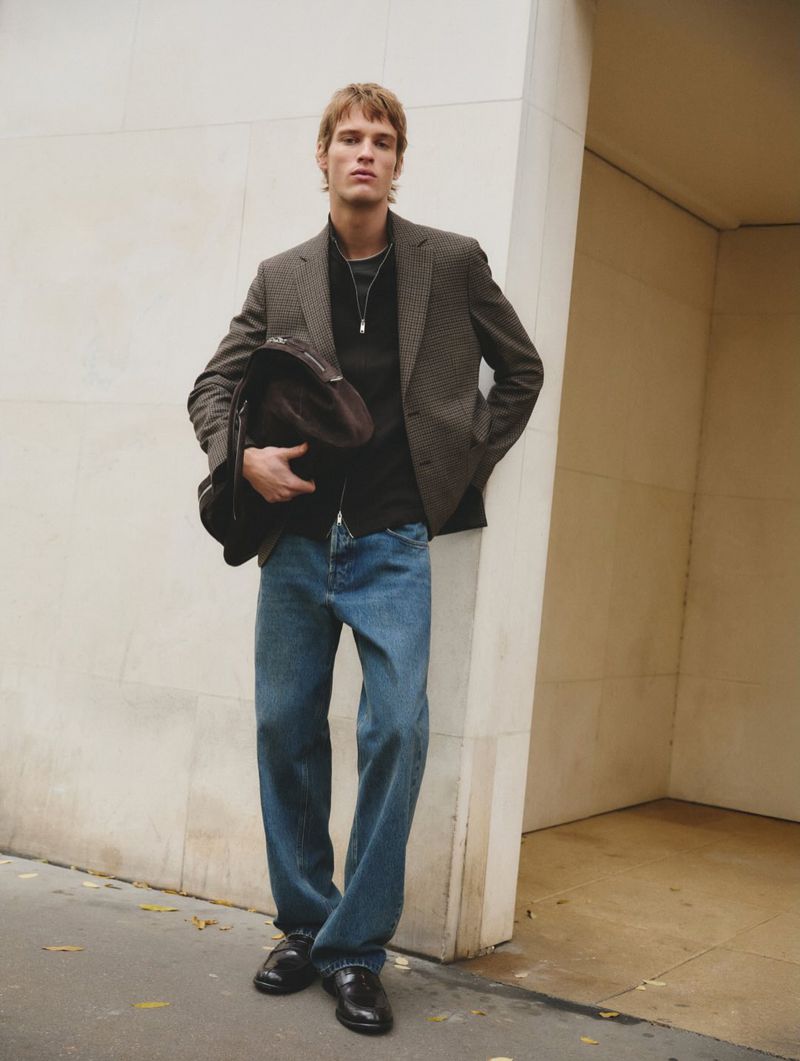 A blond male model poses against a concrete wall in a blazer, blue jeans, and black shoes, while holding a dark brown jacket.