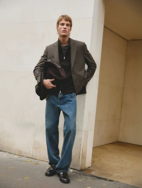 A blond male model poses against a concrete wall in a blazer, blue jeans, and black shoes, while holding a dark brown jacket.