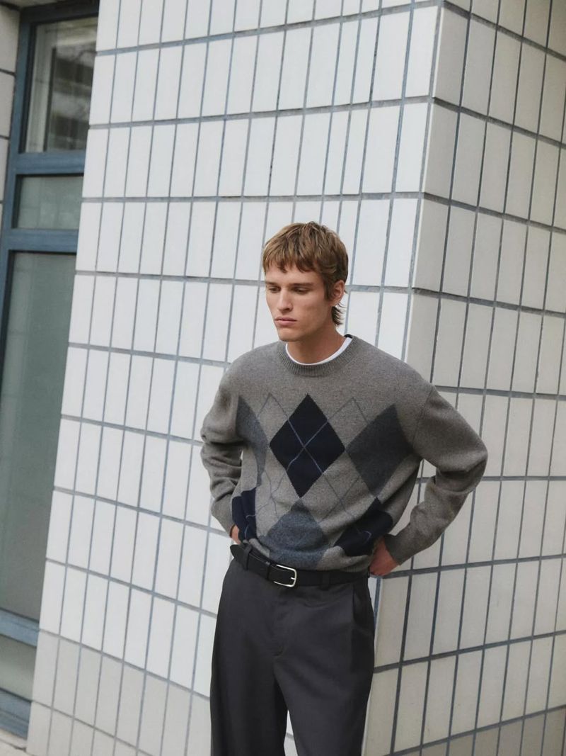 A blonde-haired man wears a gray argyle sweater, black belt, and gray pants, standing against a white tiled wall in natural light.
