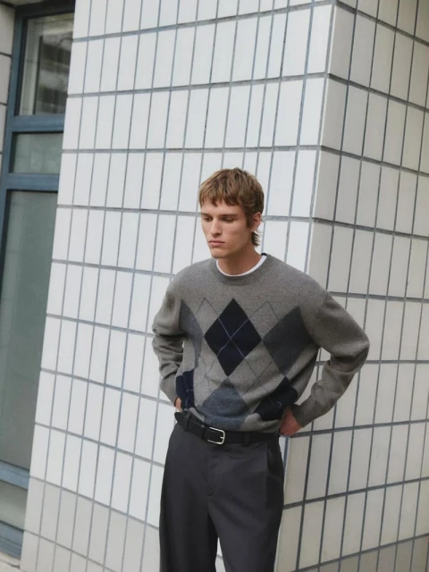 A blonde-haired man wears a gray argyle sweater, black belt, and gray pants, standing against a white tiled wall in natural light.