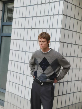 A blonde-haired man wears a gray argyle sweater, black belt, and gray pants, standing against a white tiled wall in natural light.