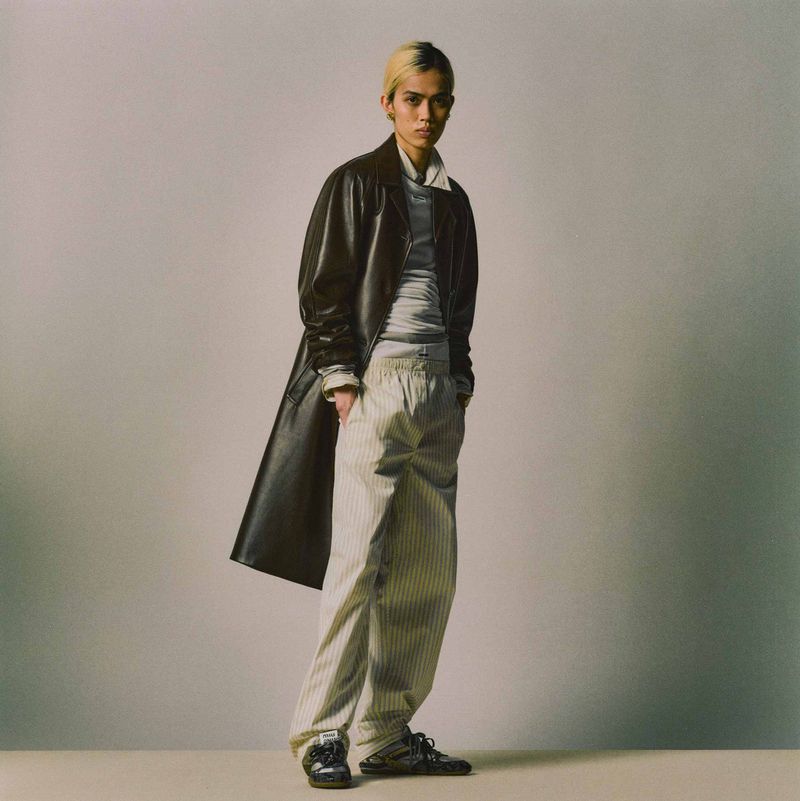 Full-length shot of a young adult with blond-streaked hair, wearing a black leather coat, light grey shirt, and striped pants in a studio.