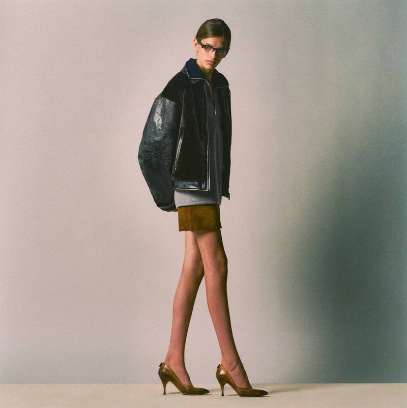 Model wearing a dark leather bomber jacket, grey sweater, and brown miniskirt with pointed-toe heels against a neutral studio background.