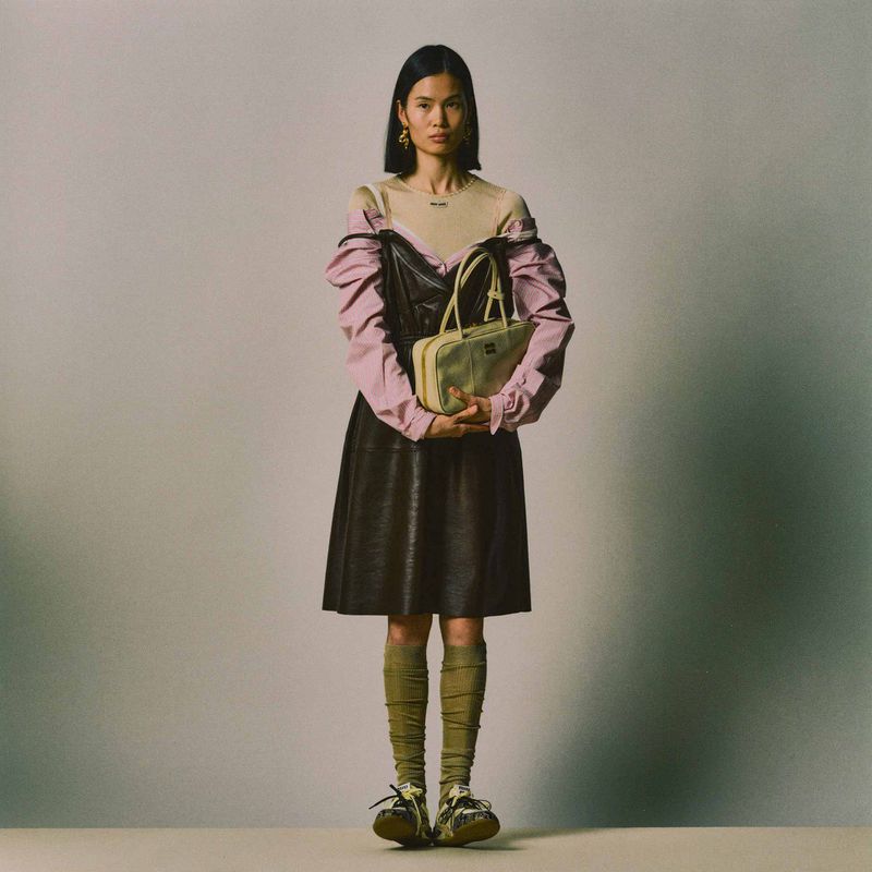 Full-length shot of an Asian woman in a dark dress and beige shirt, holding a yellow purse, standing in a neutral-toned studio.