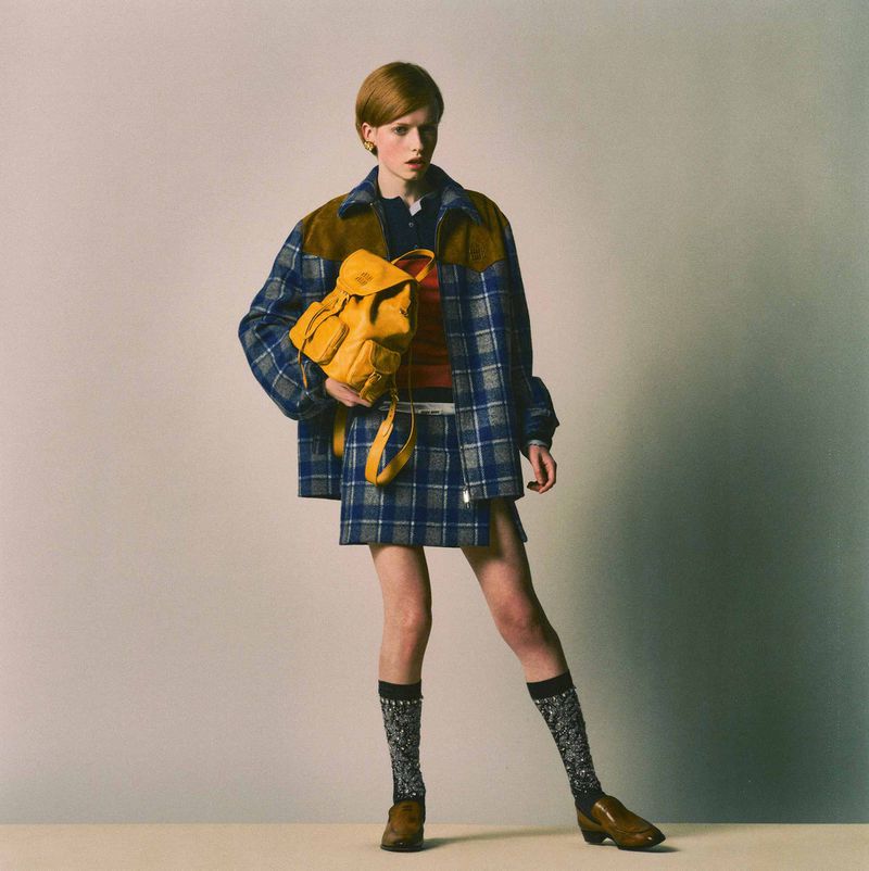 A woman poses in a blue plaid jacket, skirt, orange sweater, and knee socks, holding a yellow backpack against a neutral backdrop.