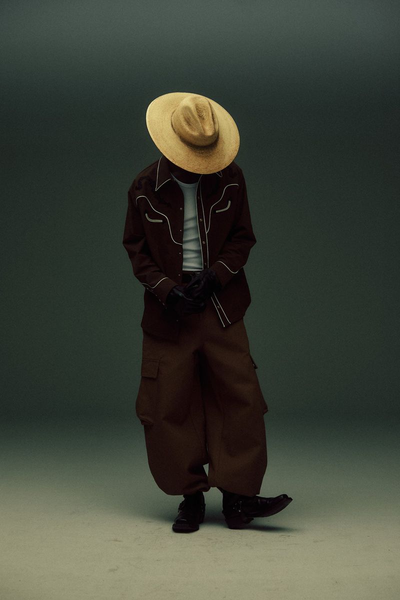 A person in a brown western shirt and baggy pants, wearing a straw cowboy hat, posing in front of a neutral gradient background.