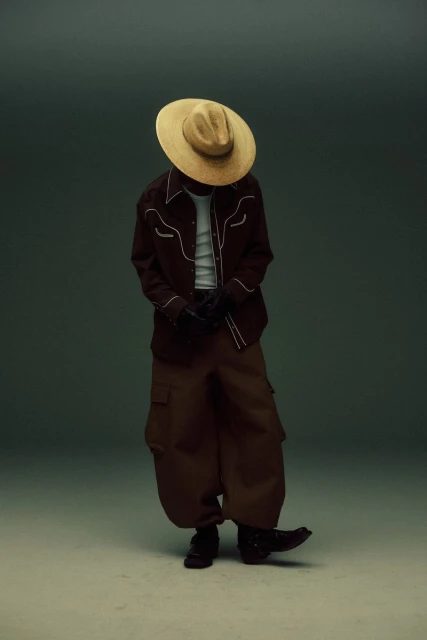 A person in a brown western shirt and baggy pants, wearing a straw cowboy hat, posing in front of a neutral gradient background.