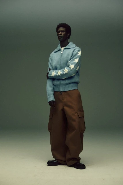 A dark-skinned male fashion model in a blue zip-up cardigan with white stars and brown cargo pants, posing in a studio with a neutral background.