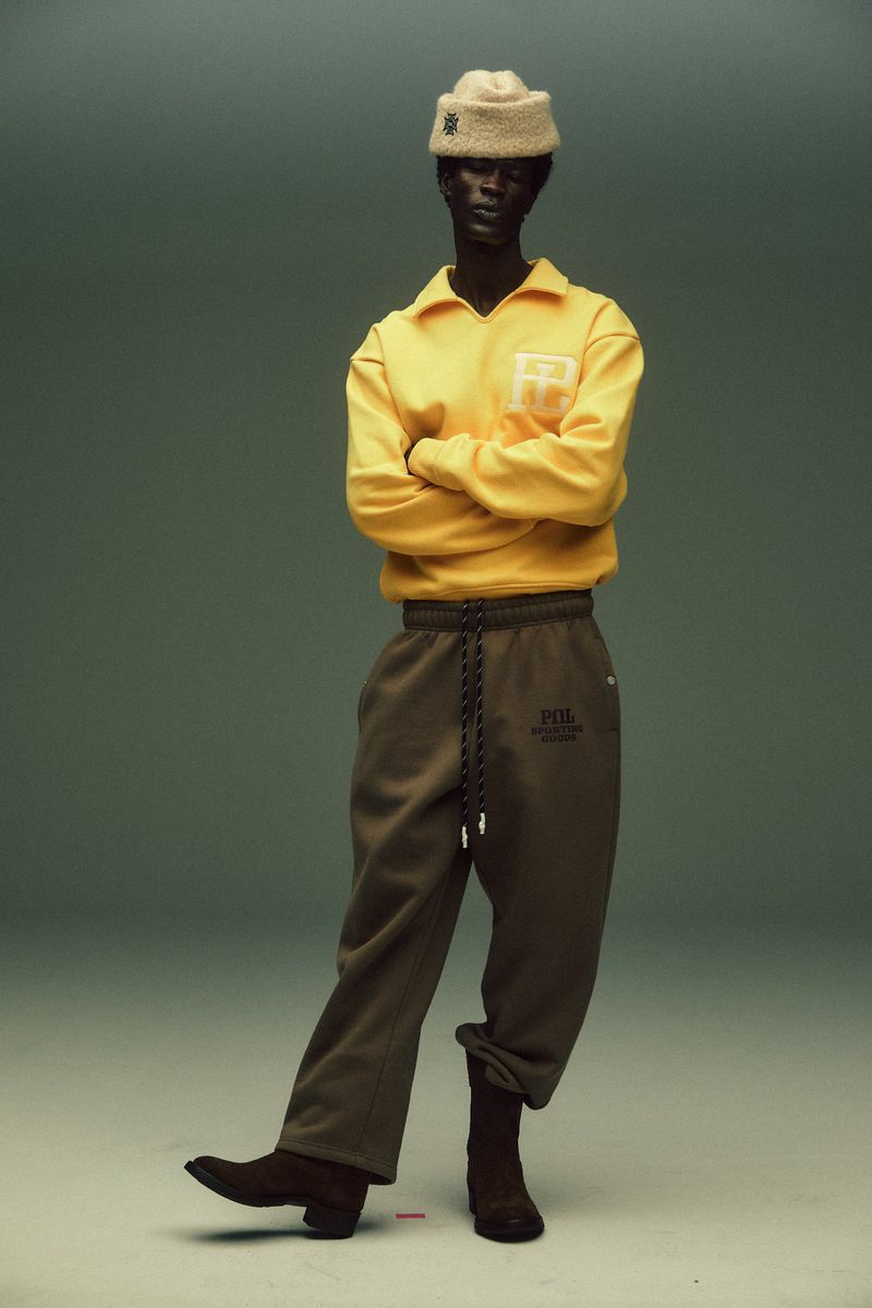Studio shot of a slender adult posing in yellow top, loose green pants, and brown boots against a muted green background.