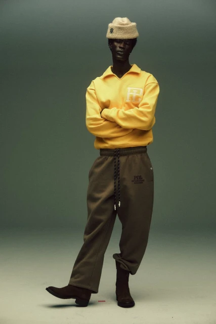 Studio shot of a slender adult posing in yellow top, loose green pants, and brown boots against a muted green background.