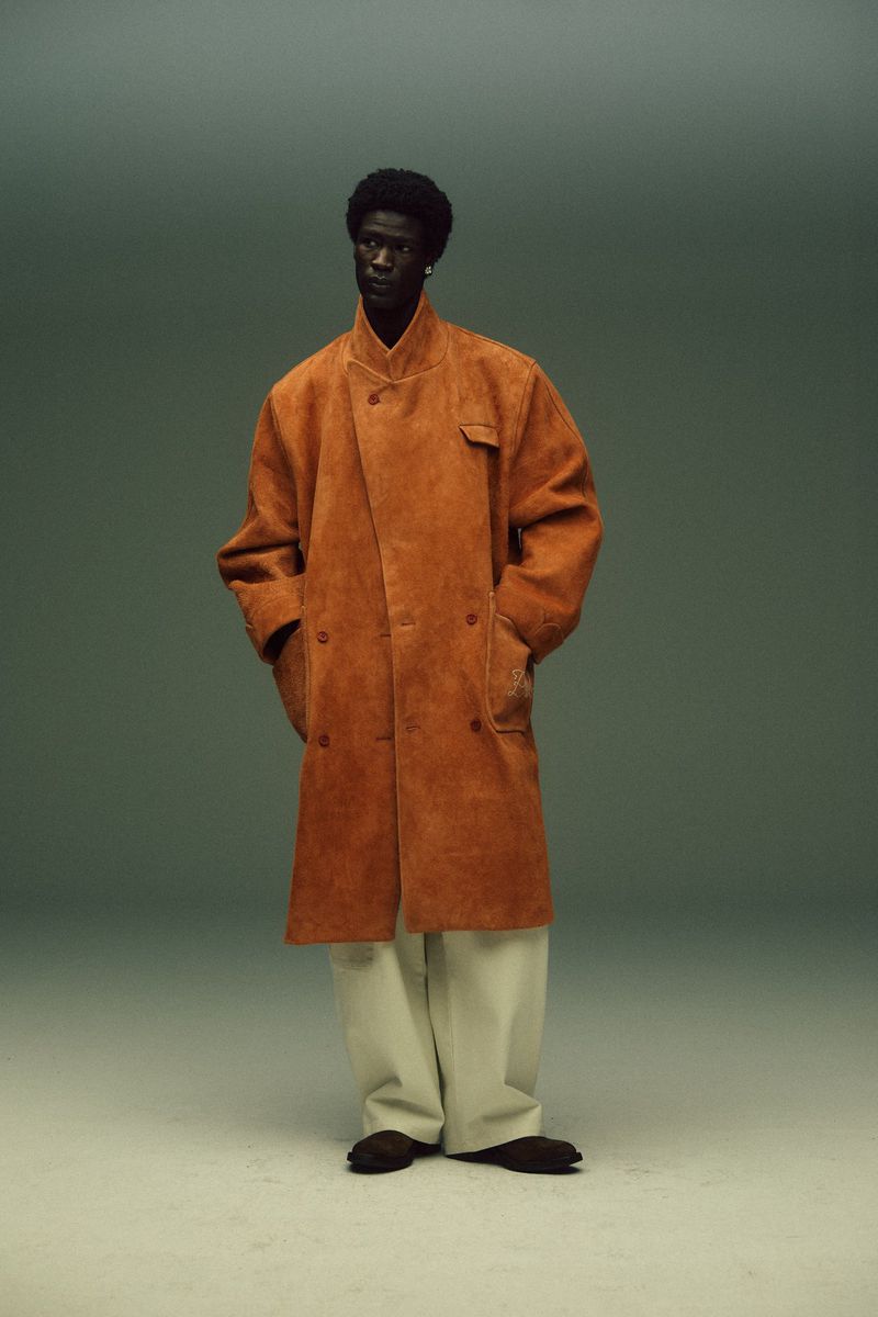 Studio shot of a Black male model in a long tan coat and cream pants, standing against a grey background, showcasing fashion.