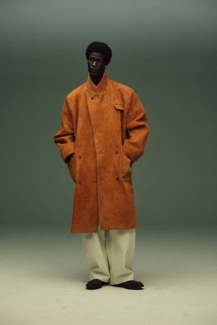 Studio shot of a Black male model in a long tan coat and cream pants, standing against a grey background, showcasing fashion.