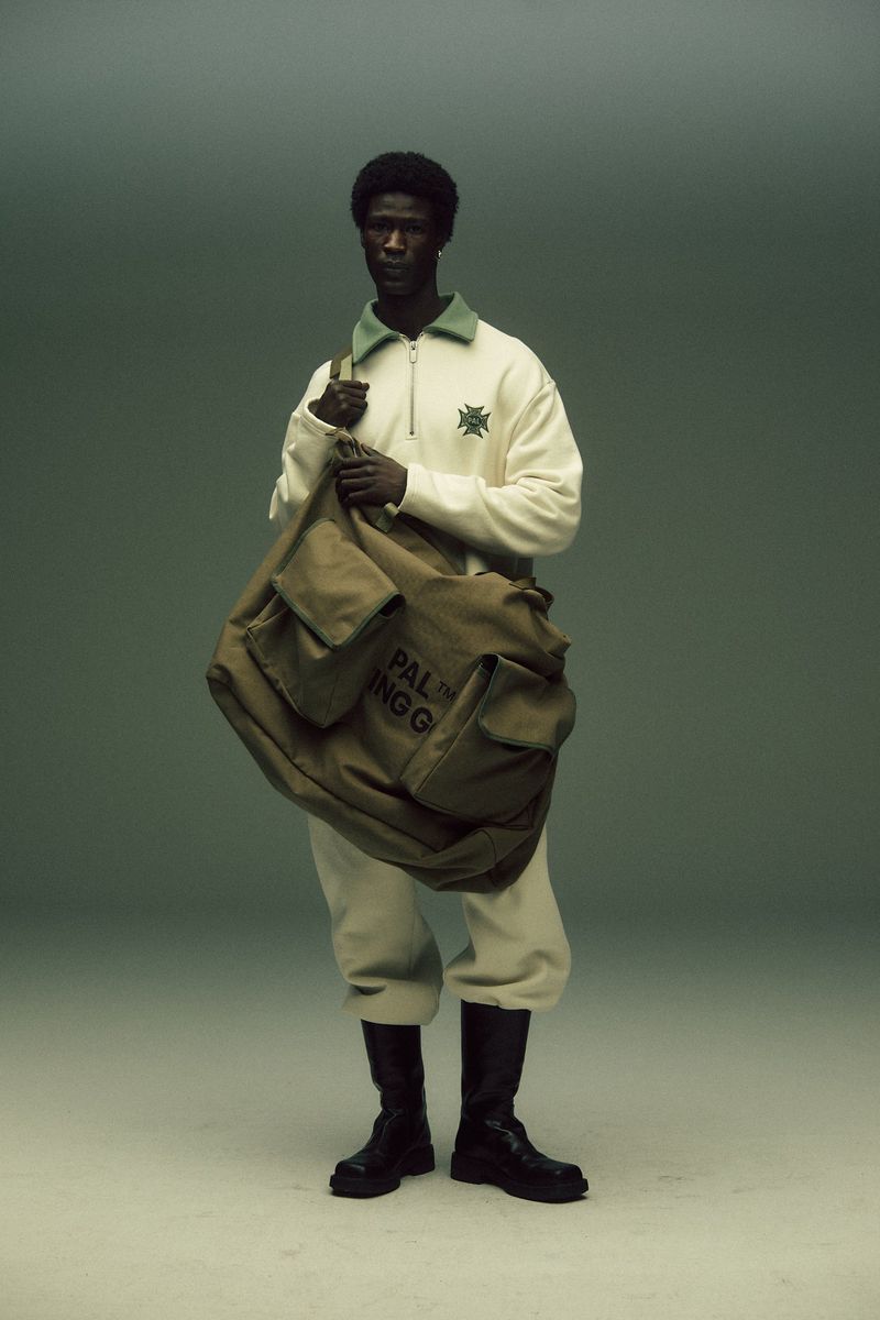A dark-skinned male fashion model poses in a photo studio wearing an off-white tracksuit, black boots, and carrying an olive duffel bag.