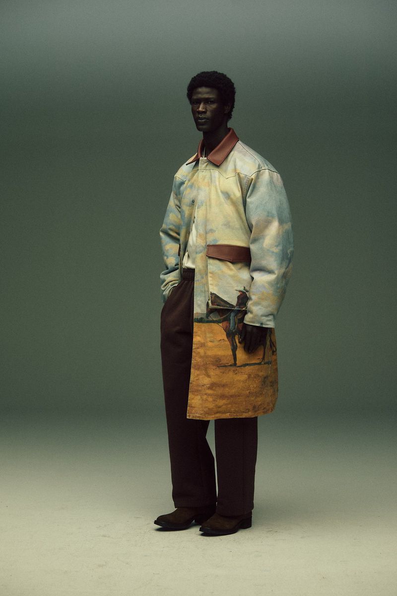 A dark-skinned man in stylish clothing with a painted design poses in a studio against a neutral background, showcasing fashion.
