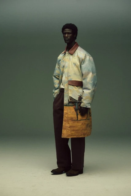 A dark-skinned man in stylish clothing with a painted design poses in a studio against a neutral background, showcasing fashion.