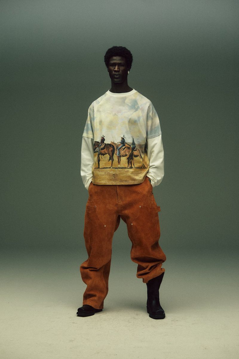 Full-length shot of an adult male model with dark skin and curly hair wearing a printed long-sleeve shirt, orange pants, and black boots.