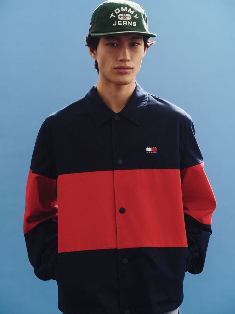 Portrait of a young adult male wearing a green Tommy Jeans cap and a color-blocked, red and navy jacket against a light blue backdrop.