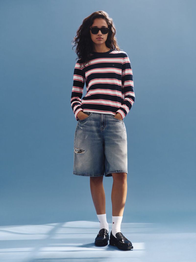 Woman in a striped sweater and denim shorts poses in front of a blue backdrop, accented by subtle lighting effects on the floor.