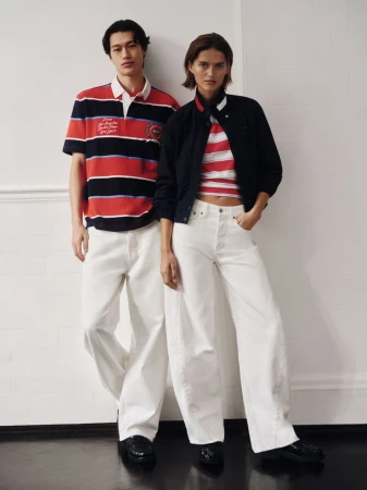 Two fashion models in striped tops and white pants pose against a wall in a fashion editorial shot with a neutral color palette.