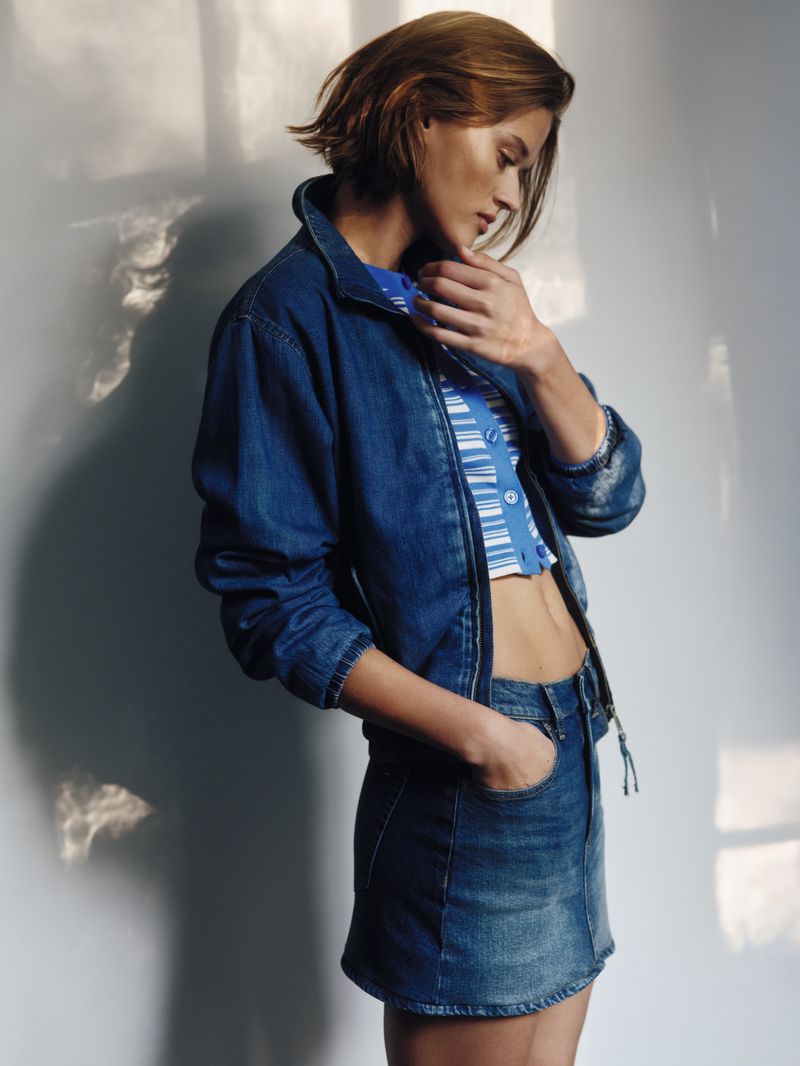 A young woman models a denim jacket and mini skirt, posing indoors with soft, diffused light casting shadows on the wall behind her.